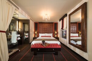 Habitación de hotel con una cama grande, un banco tapizado en rojo, un espejo amplio y una araña.