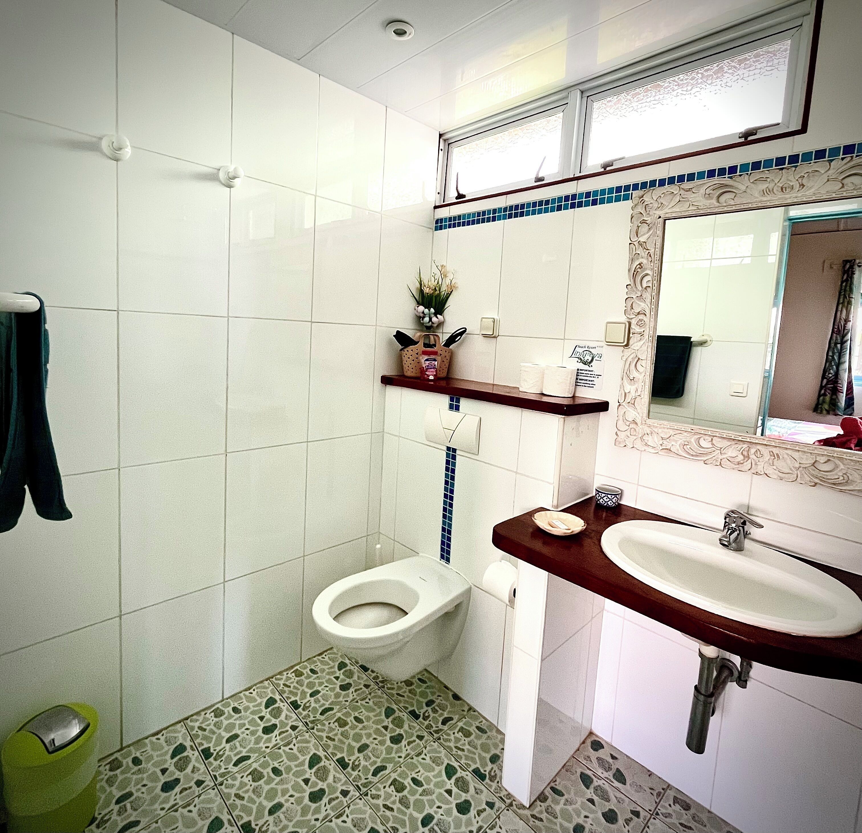 A bathroom with a toilet, sink, and mirror. There is a towel rack and a trash bin.