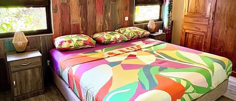 Un dormitorio con cabecera de madera, colcha colorida, mesita de noche con lámpara y armario de madera.
