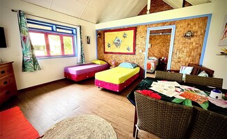 Una habitación con dos camas, piso de madera, un ventanal con cortinas, pared con diseño tejido y una silla de mimbre.