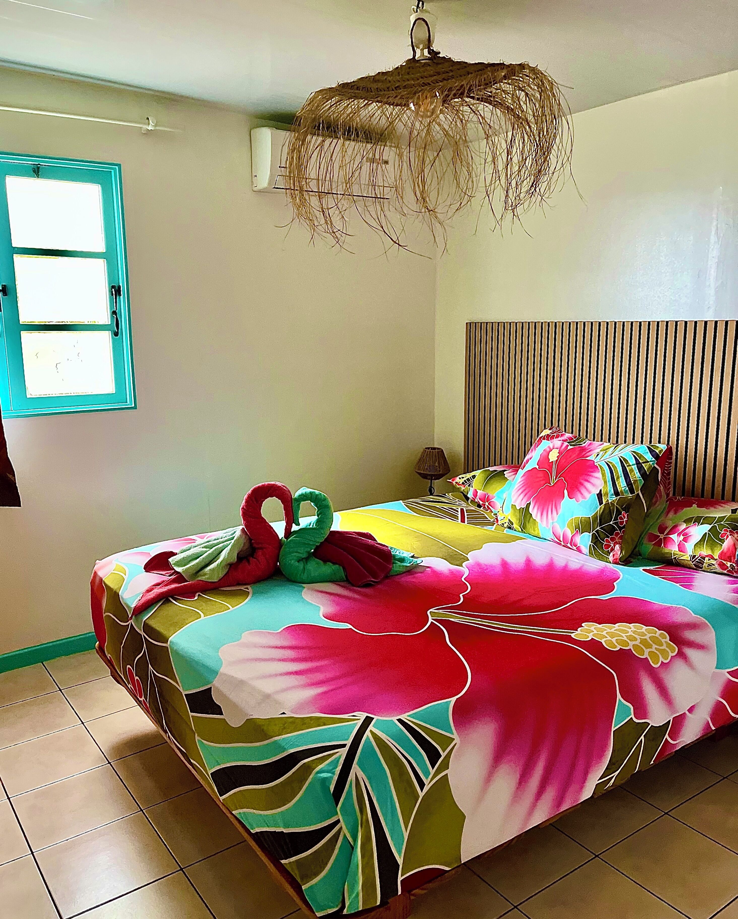 A bed with a floral bedspread, a hanging light fixture, a window with blinds, and a wall-mounted air conditioner.