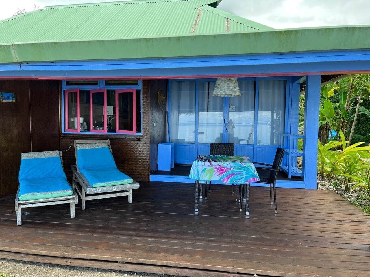 A wooden building with a blue exterior and a covered porch. There are two lounge chairs with blue cushions and a table with a colorful tablecloth.