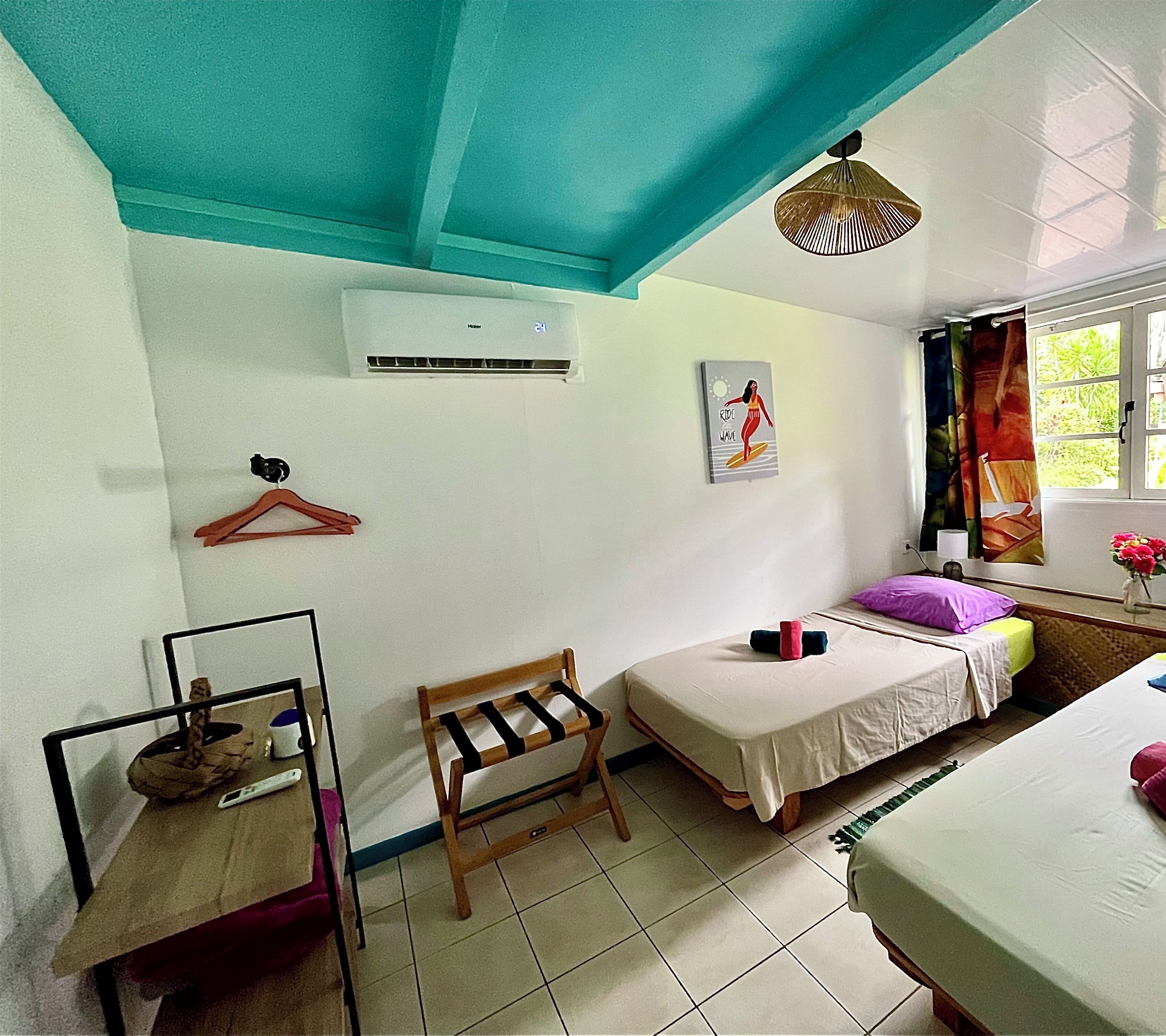 A room with a bed, a chair, a small table, a wall-mounted air conditioner, a window with curtains, and a wall with a picture.