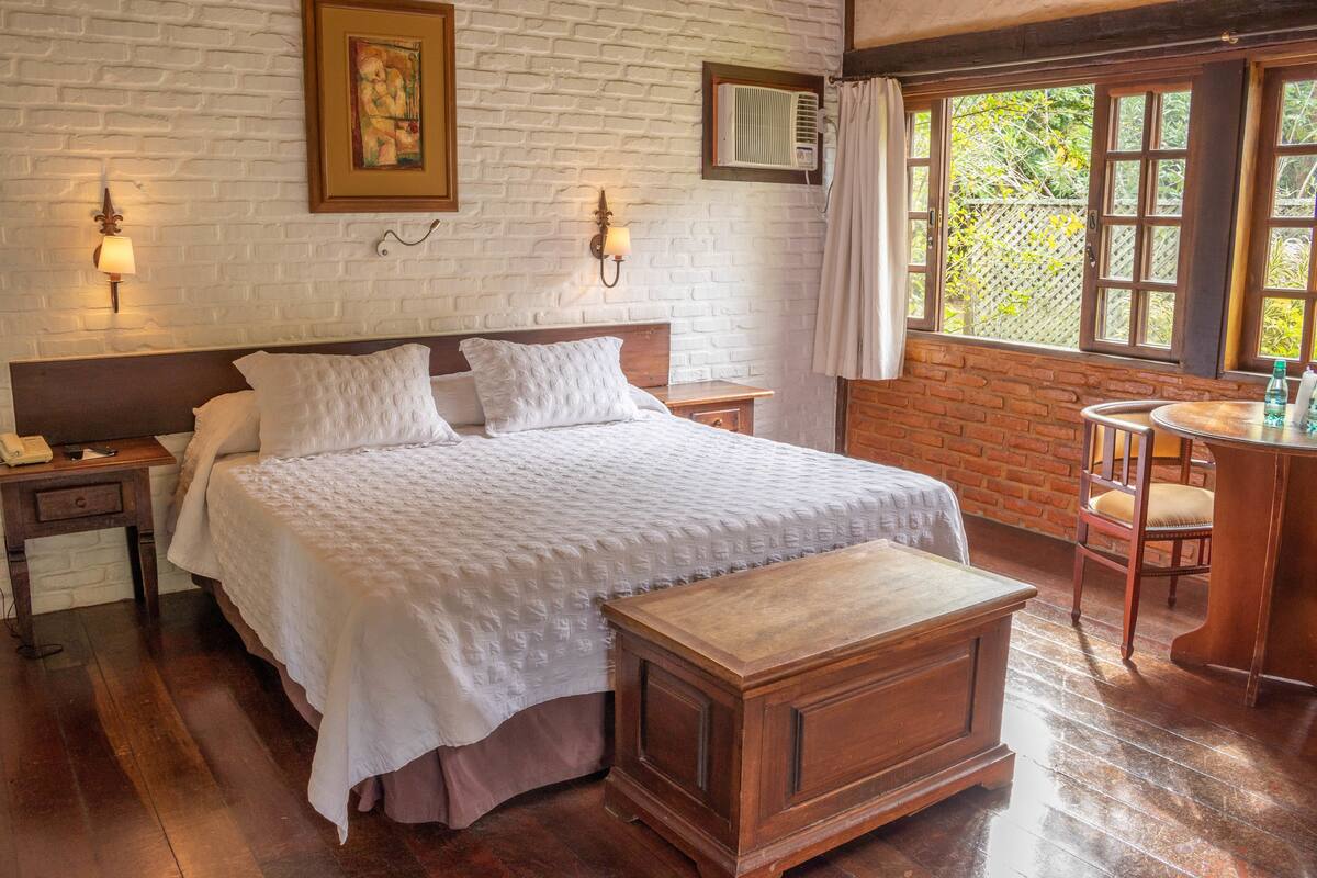 A bedroom with a large bed, wooden nightstands, a wooden bench, a small table, and a window with a view of greenery.