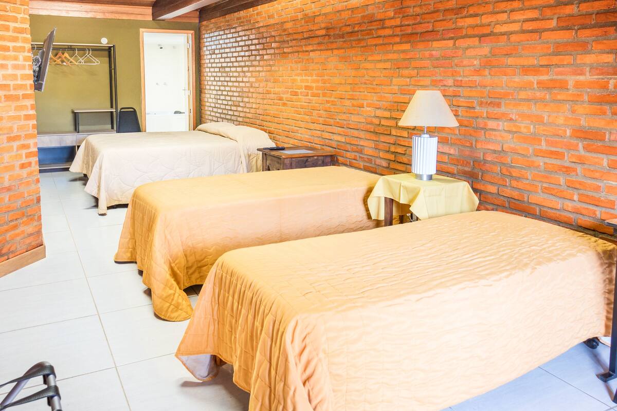 A hotel room with three beds, a brick wall, a door, a table with a lamp, and a bicycle.