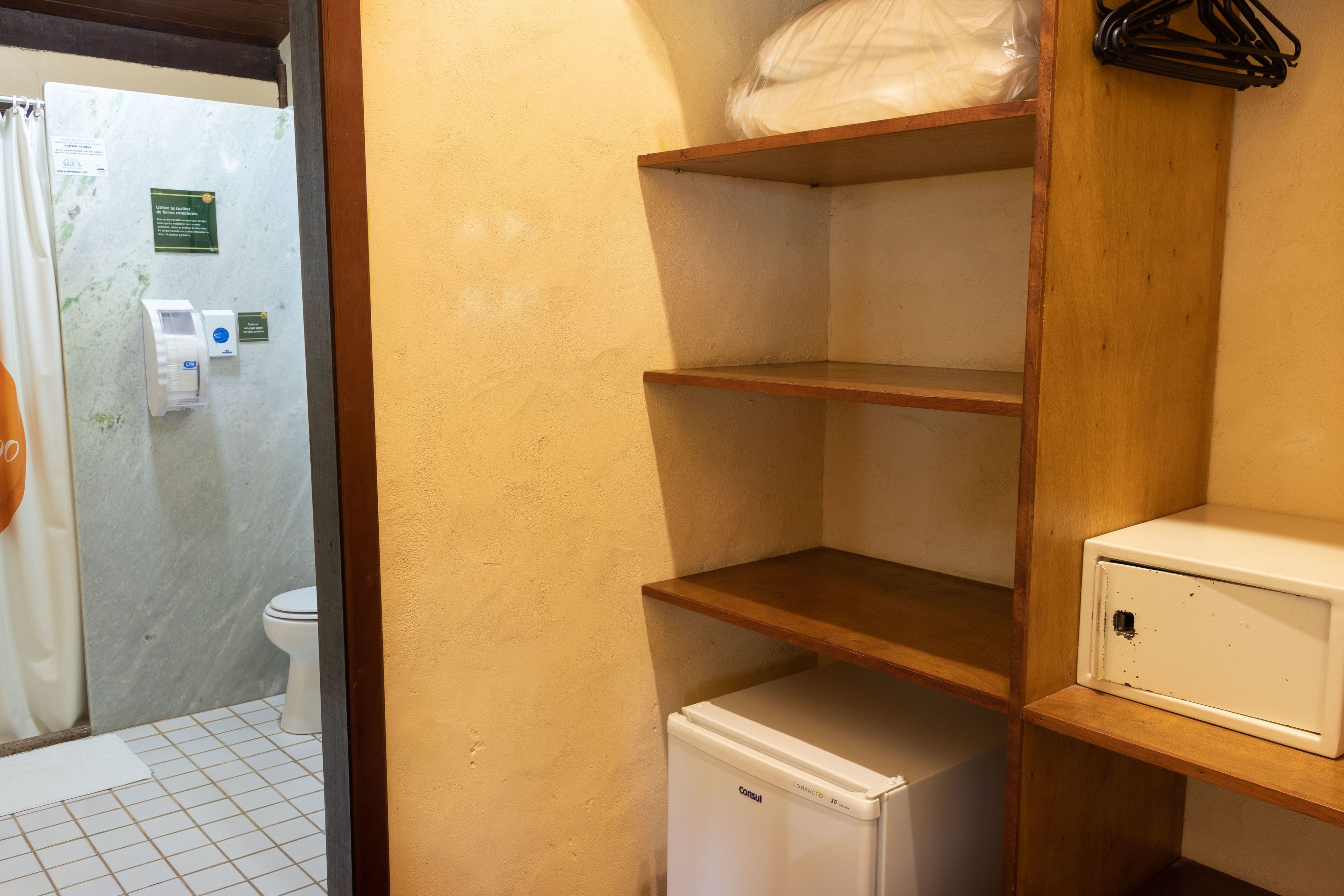 A bathroom with a toilet, a shower curtain, a small refrigerator, and a shelf with a plastic bag.