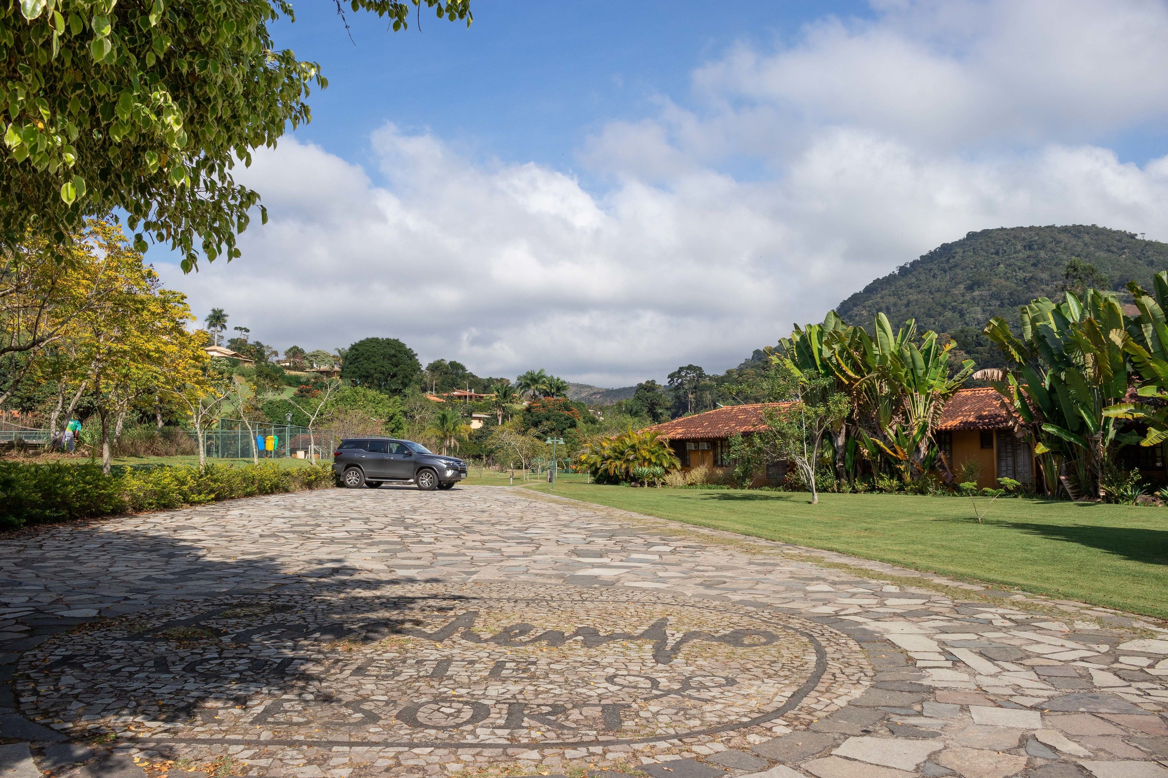 A paved driveway leading to a building with a tiled roof, surrounded by lush greenery and a mountain in the background.