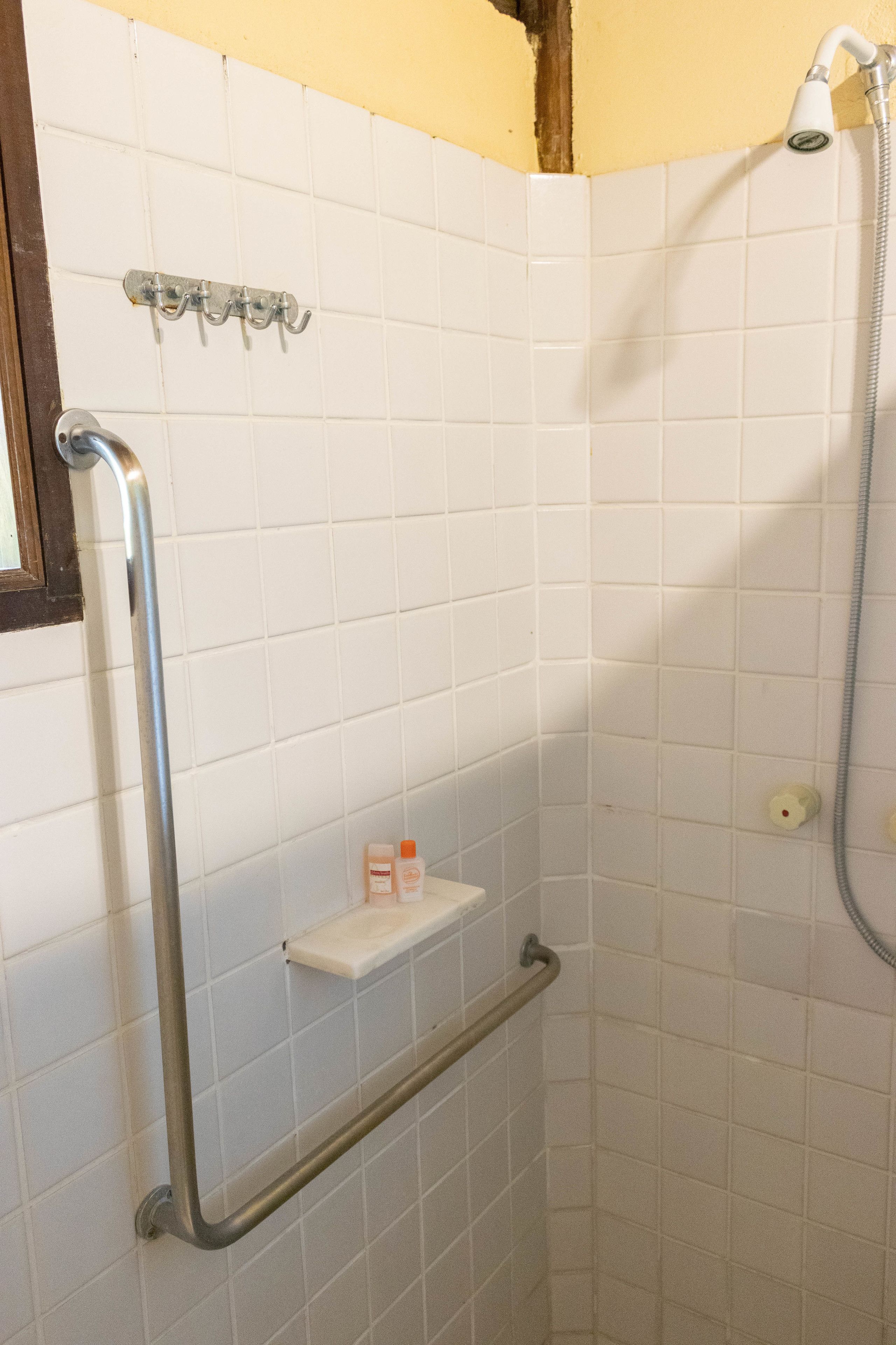 A corner bathroom with white tiles, a shower, a grab bar, and a shelf with toiletries.