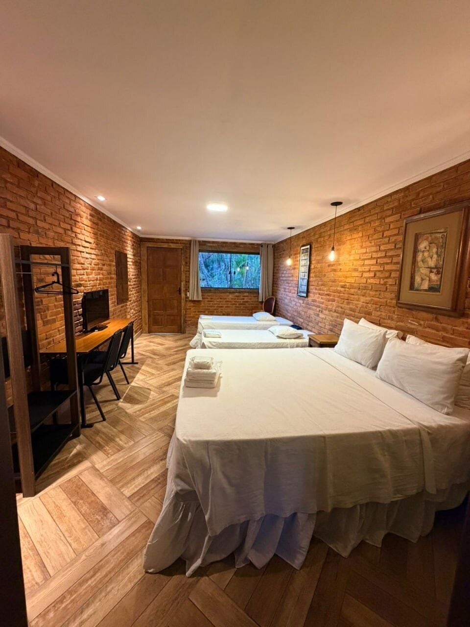 A room with a brick wall, a wooden desk, two beds, and a window with a view.