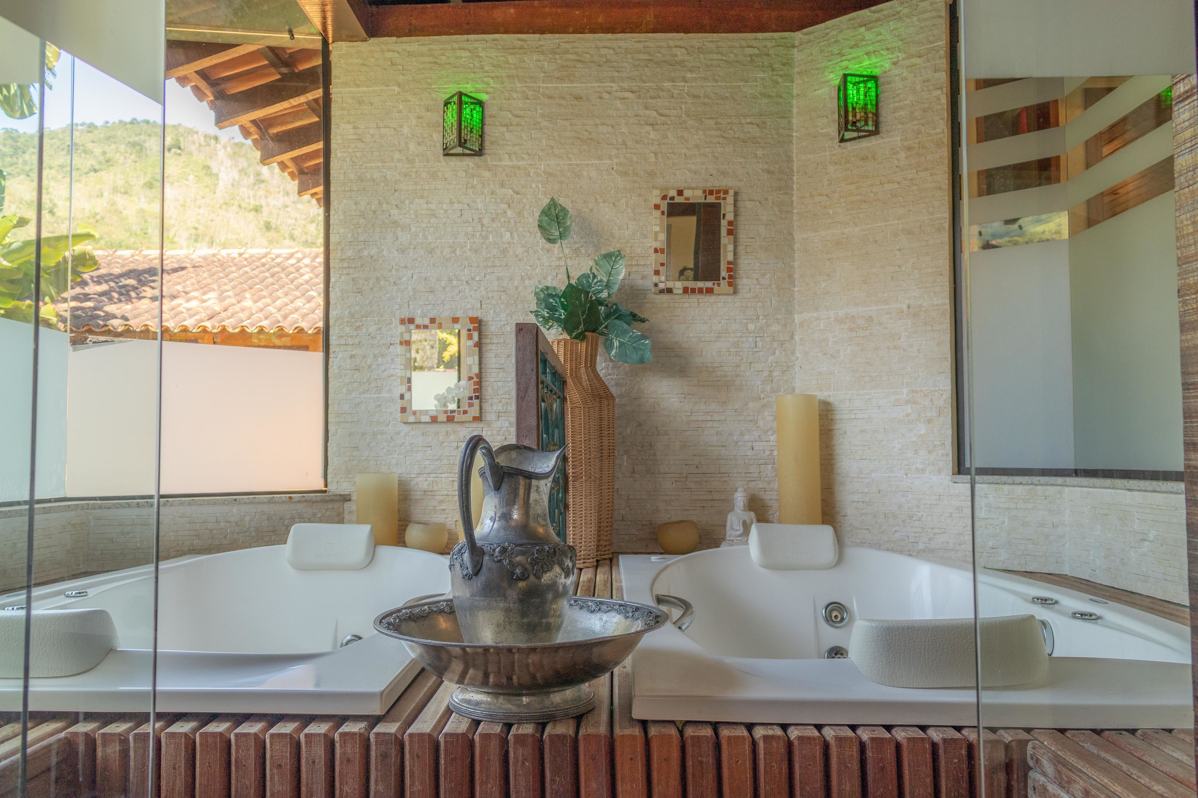 A bathroom with two large white tubs, a decorative pitcher, a mirror, and a brick wall.