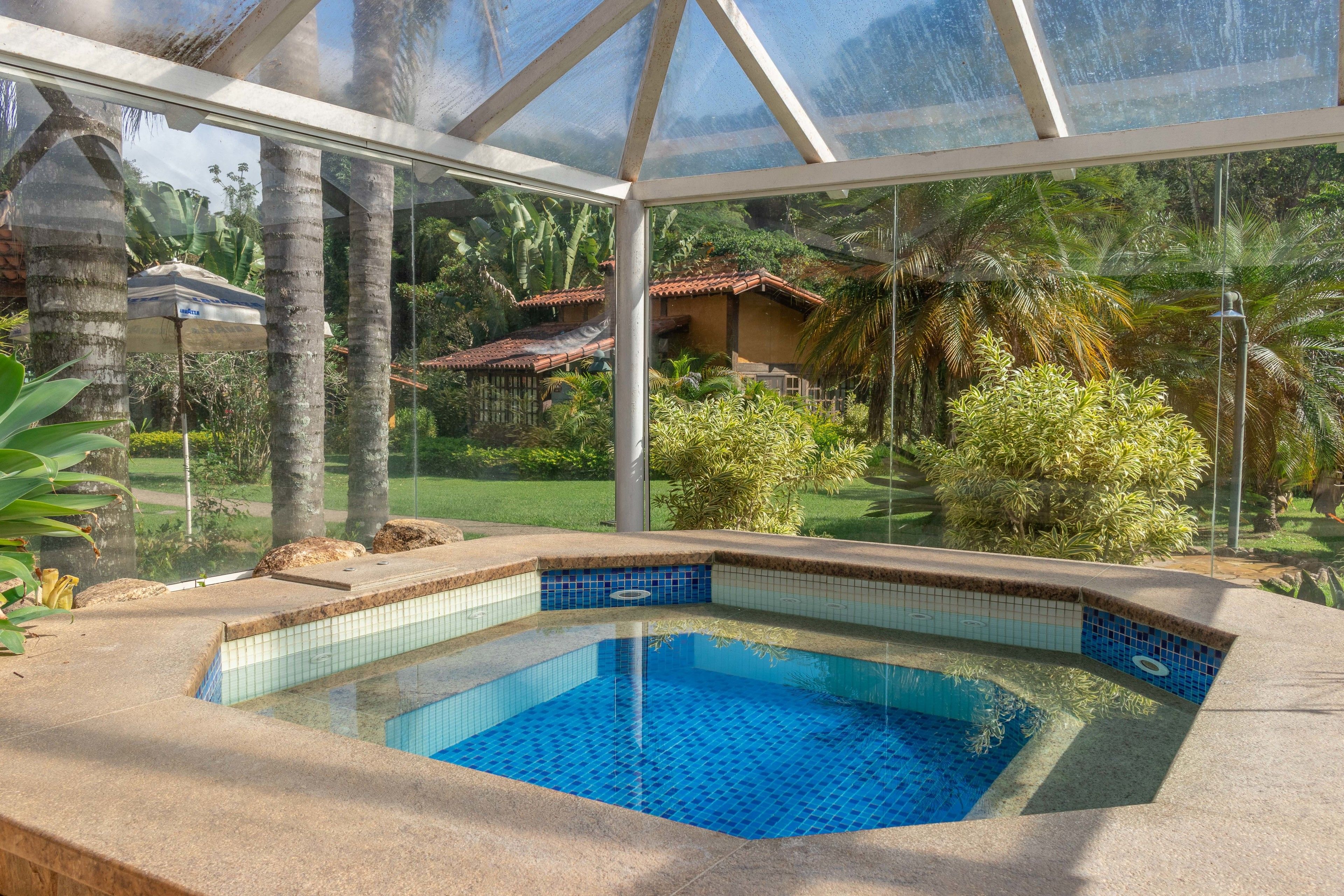 A glass-enclosed swimming pool with a tiled edge, surrounded by tropical plants and a view of a residential area.