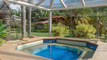 A glass-enclosed swimming pool with a tiled edge, surrounded by tropical plants and a view of a residential area.