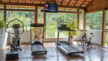 A covered gym area with exercise equipment, large windows, and a TV screen displaying a program.