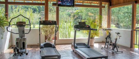A covered gym area with exercise equipment, large windows, and a TV screen displaying a program.