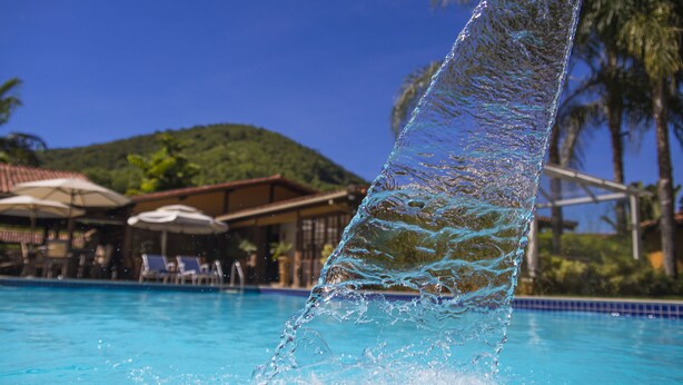 A clear view of a swimming pool with water cascading over the edge, surrounded by lounge chairs and umbrellas. - Hotel Bomtempo Itaipava (Petrópolis)