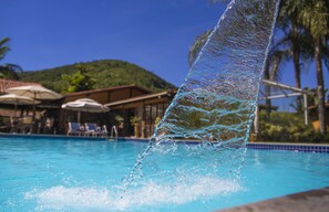 Vista despejada de una piscina con agua cayendo por el borde, rodeada de sillas de descanso y sombrillas.