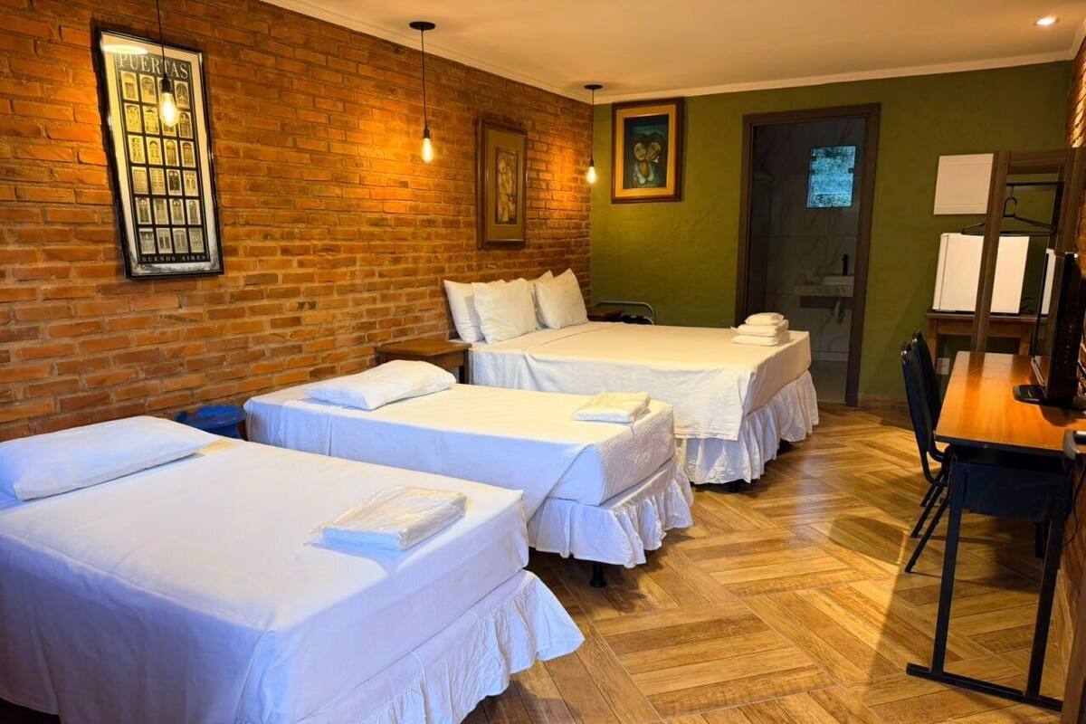 A hotel room with three beds, a wooden floor, a brick wall, and a framed picture.