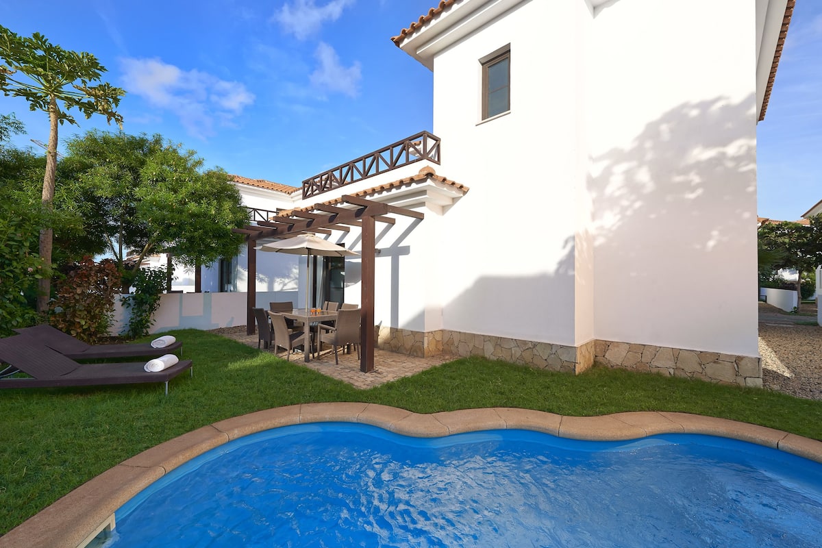 A swimming pool surrounded by a well-maintained lawn, a pergola with outdoor furniture, and a white building with a tiled roof.