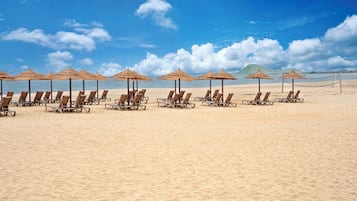 Ein Strand mit Liegestühlen, reetgedeckten Sonnenschirmen, einem Volleyballnetz und blauem Himmel.