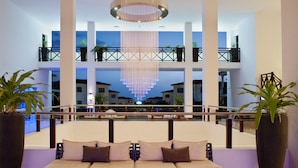 Una hall d'hotel moderna con un'area salotto centrale, un lampadario grande e vista sull'interno dell'edificio.
