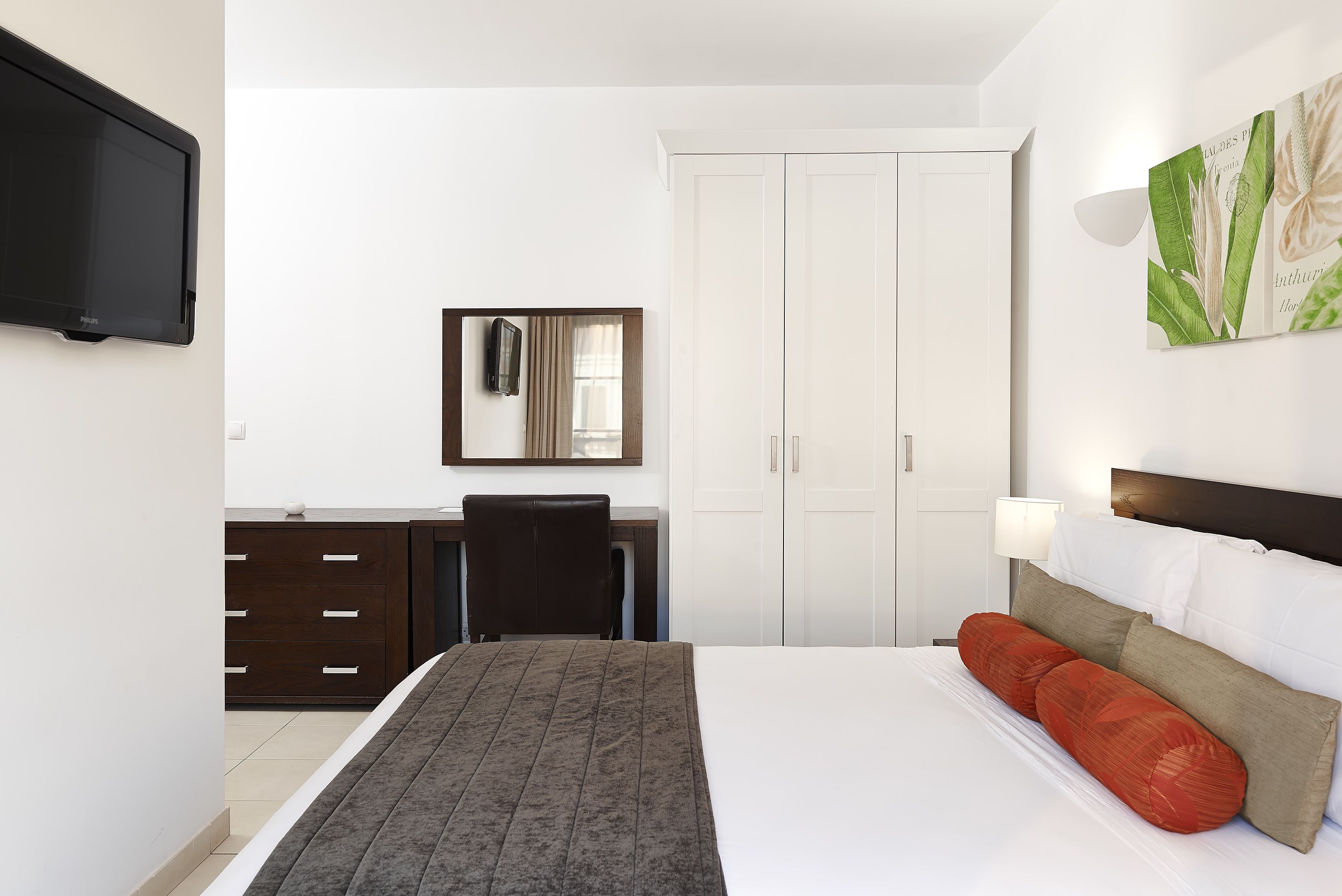 A hotel room with a bed, a desk, a chair, a wardrobe, and a flat-screen TV mounted on the wall.