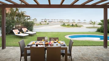Un área de comedor al aire libre con una mesa lista para el desayuno, con vista a una piscina y una playa.