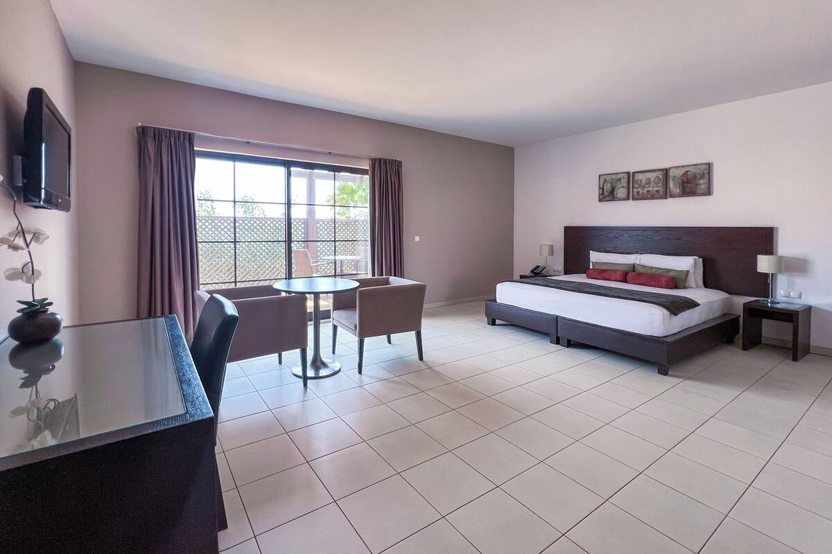 A hotel room with a bed, a desk, and a chair. There is a window with curtains, a TV mounted on the wall, and a small table with a lamp.