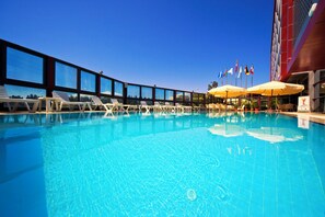 A swimming pool with lounge chairs and umbrellas, surrounded by large windows and flags. - Volley Hotel Istanbul (Istanbul)
