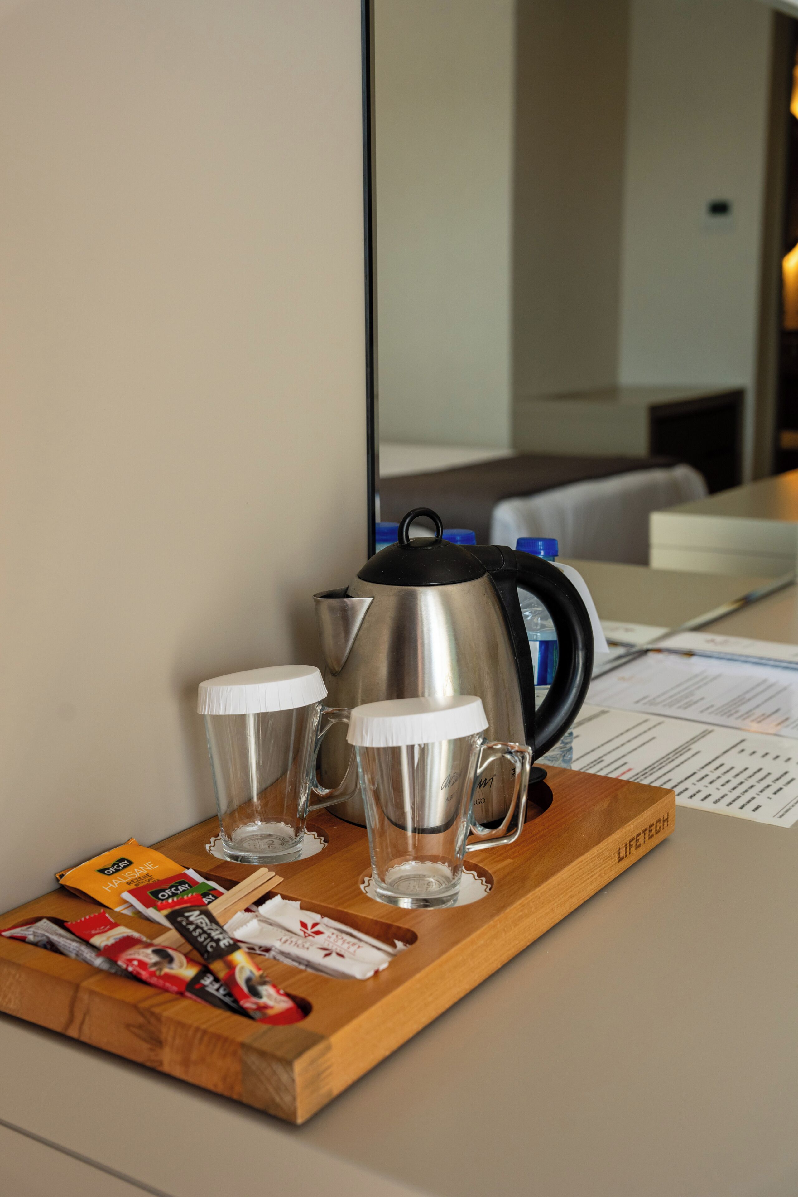 A wooden tray with a stainless steel coffee pot, two clear glasses, and some packaged snacks on a hotel table.