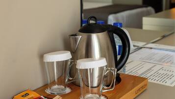 A wooden tray with a stainless steel coffee pot, two clear glasses, and some packaged snacks on a hotel table.