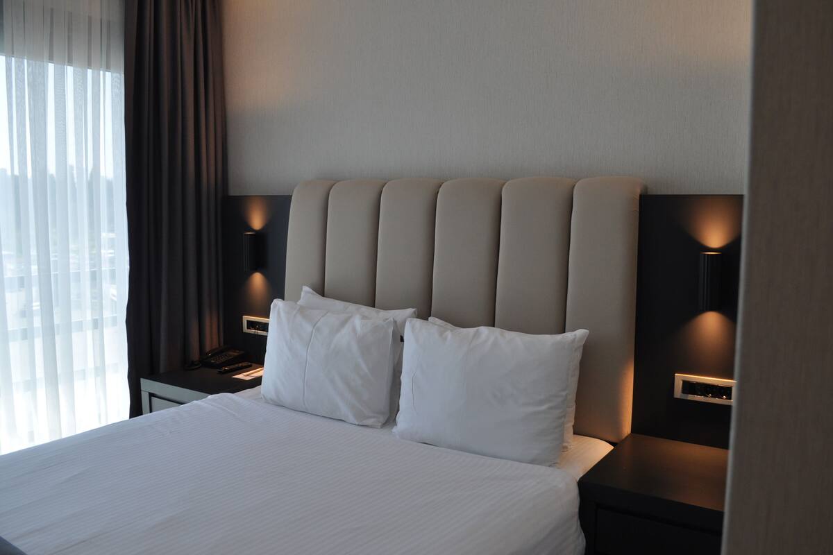 A hotel room with a large bed, two bedside tables, a headboard, and a window with curtains.