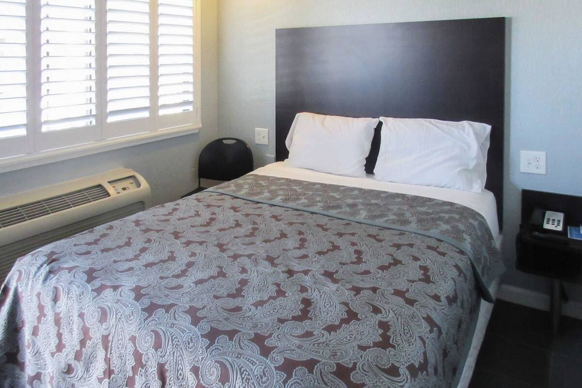 A hotel room with a bed, a heater, a window with blinds, and a nightstand with a phone.