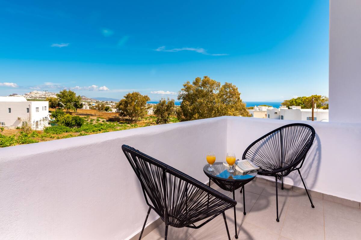 A balcony with two black chairs and a small table, overlooking a coastal landscape.