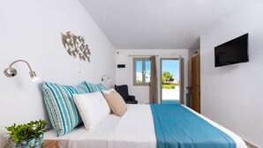 A bedroom with a bed, bedside table, lamp, TV, and a view of the outdoors. - Hotel Kalisperis (Santorini)