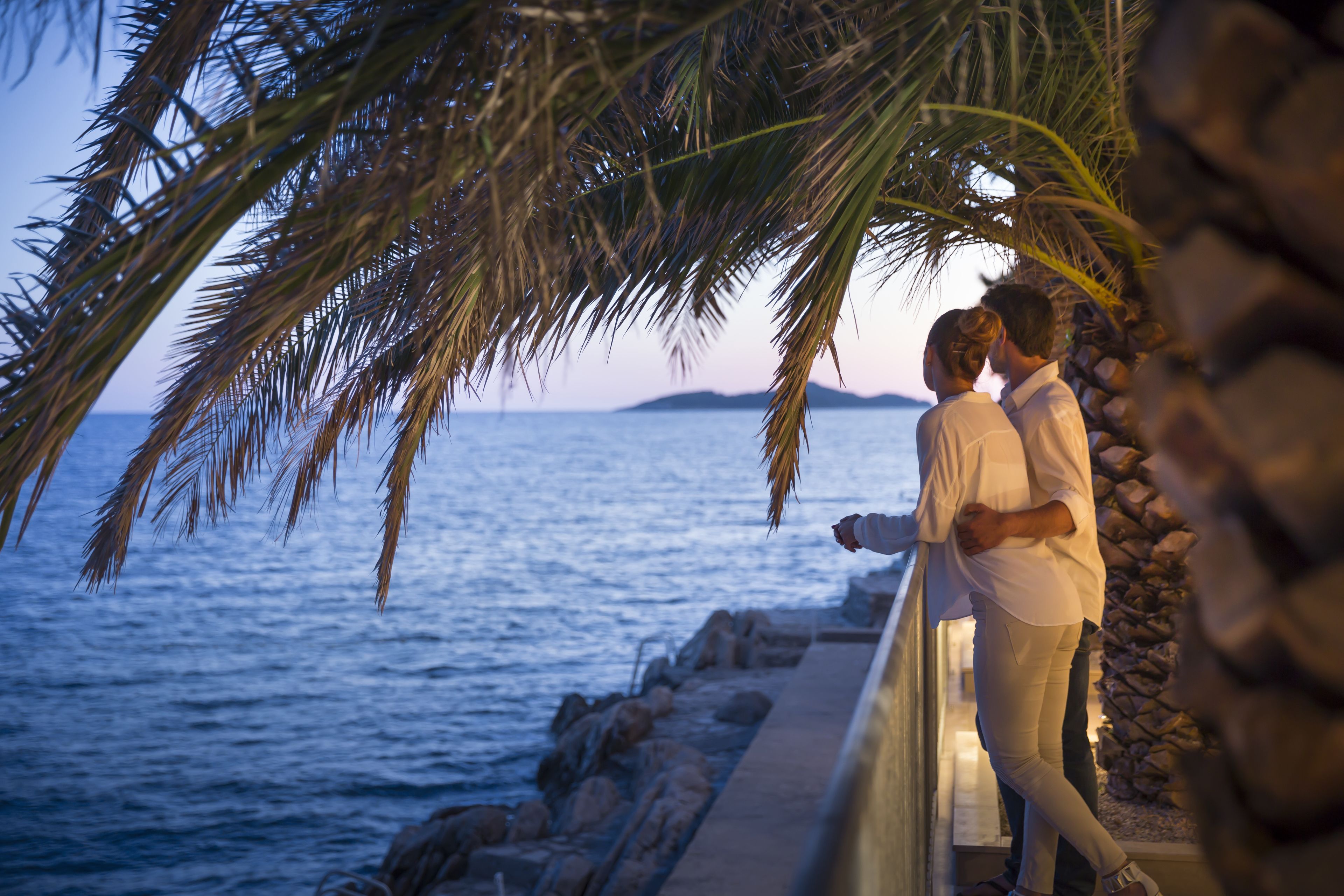 A couple standing by a seaside walkway with a view of the ocean and distant land.