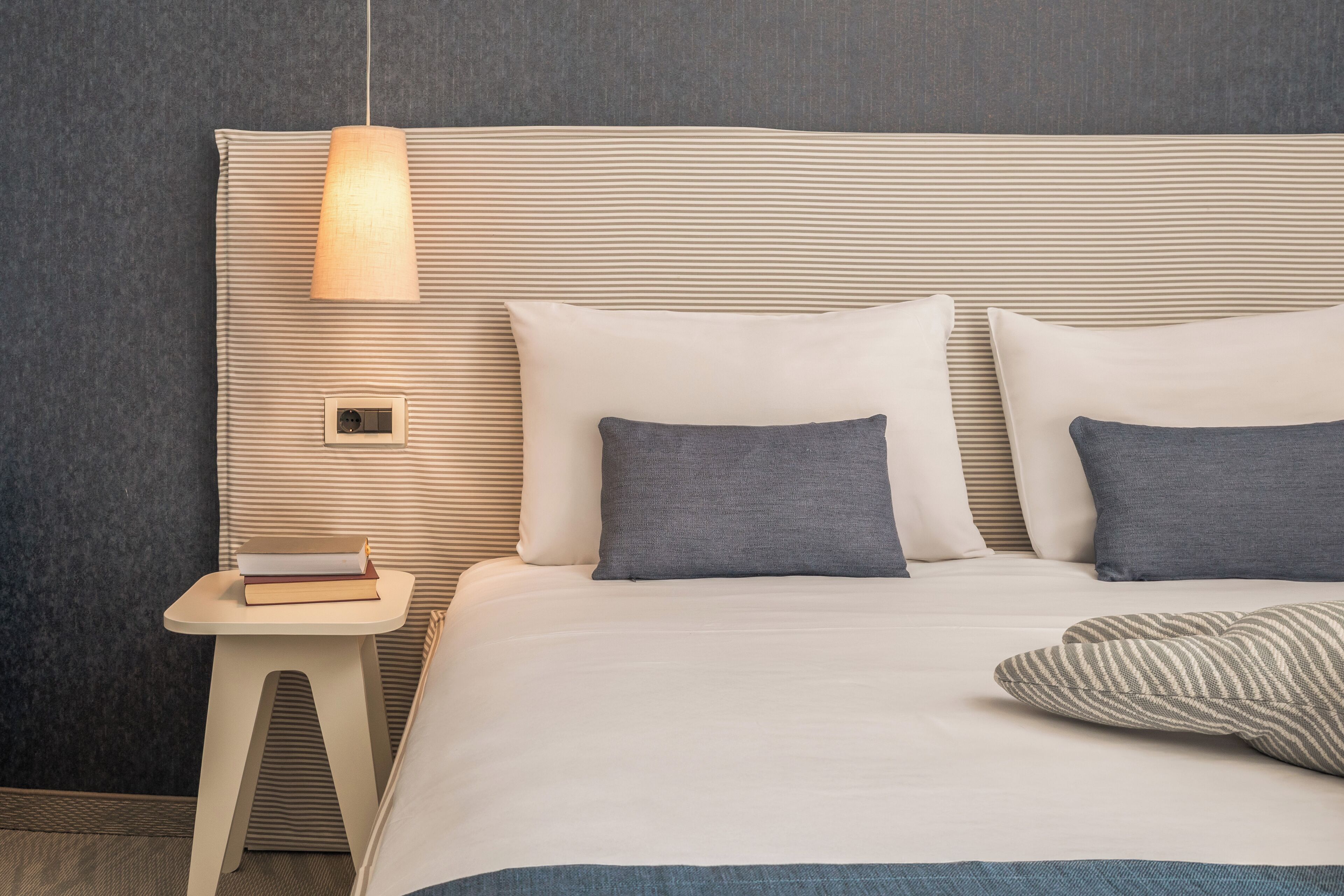 A neatly made bed with white and gray bedding, a small bedside table with a lamp, and a wall-mounted light fixture.