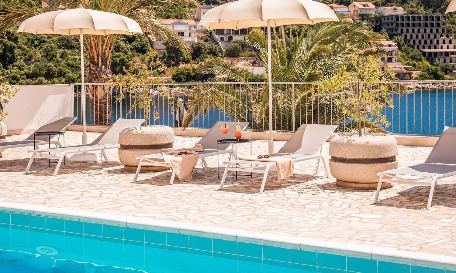 A poolside area with lounge chairs, umbrellas, and a view of the sea and buildings.