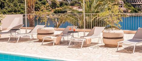 A poolside area with lounge chairs, umbrellas, and a view of the sea and buildings.