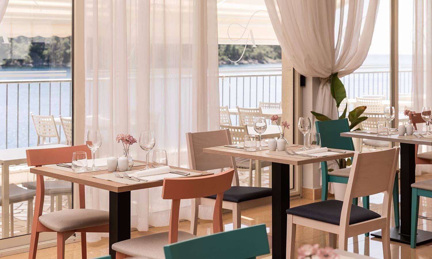A restaurant with tables set for dining, chairs, and a view of the sea.