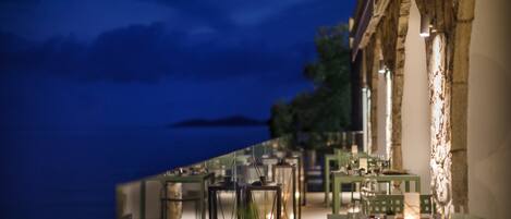 An outdoor dining area with tables and chairs, a view of the sea, and a stone building with a light fixture.