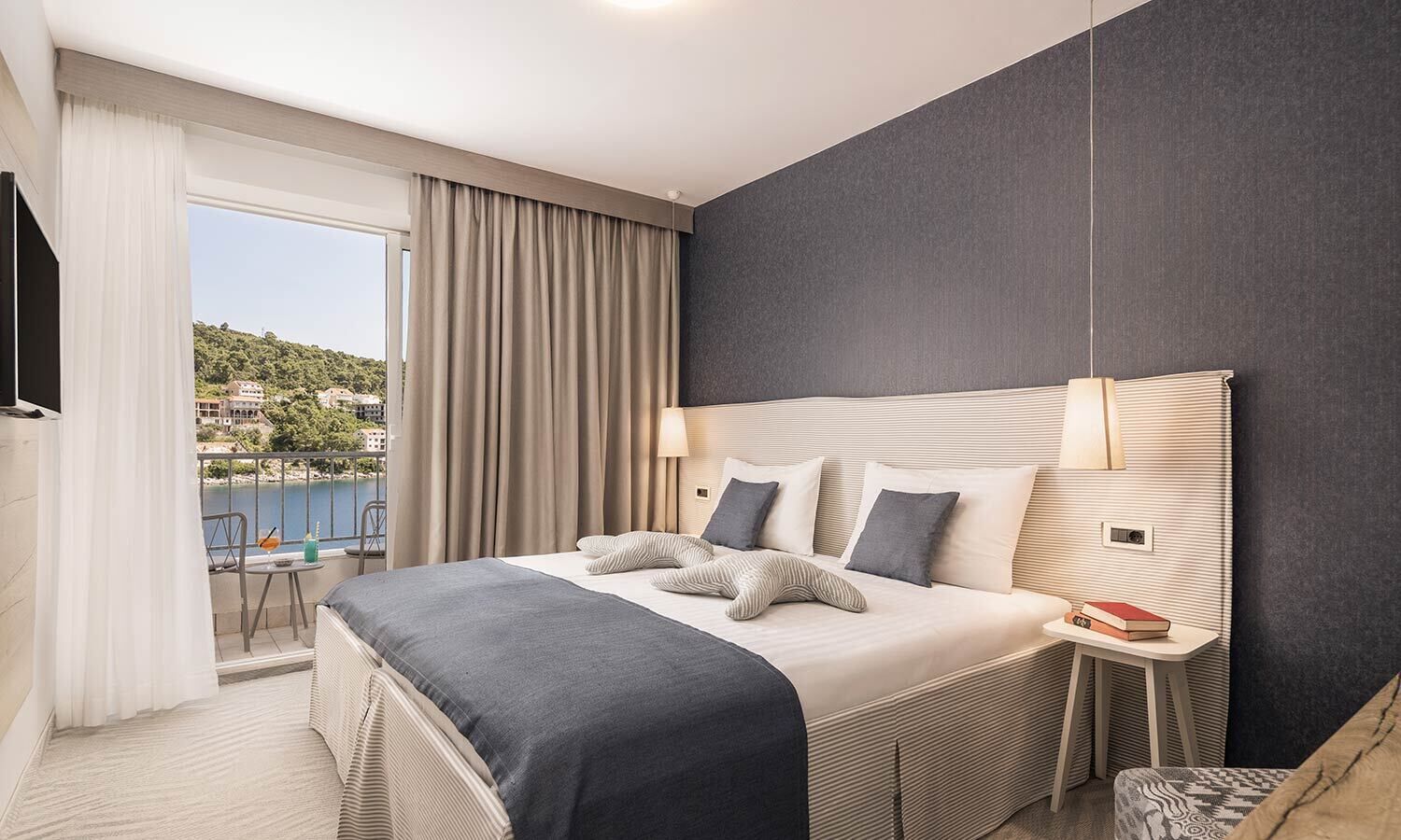 A modern hotel room with a large bed, a bedside table, a chair, and a view of the sea.
