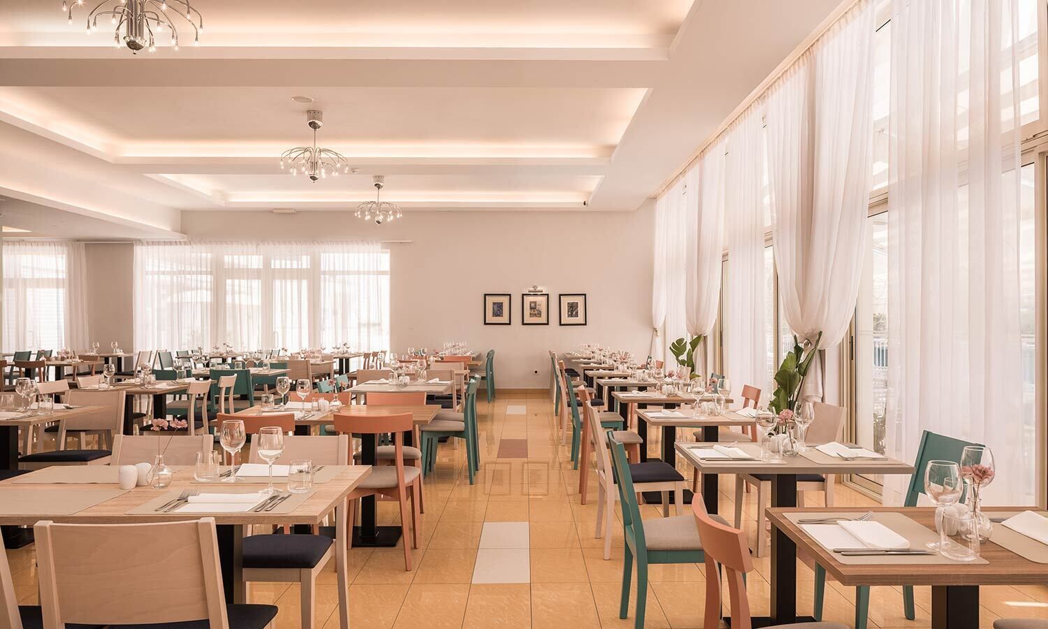 A spacious dining area with multiple tables and chairs, large windows with sheer curtains, and a chandelier.