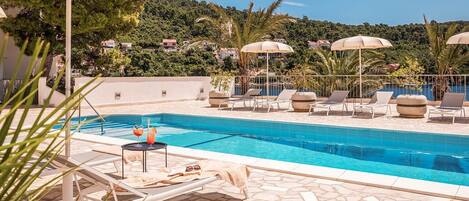 A swimming pool with lounge chairs and umbrellas, surrounded by a stone patio and lush greenery.