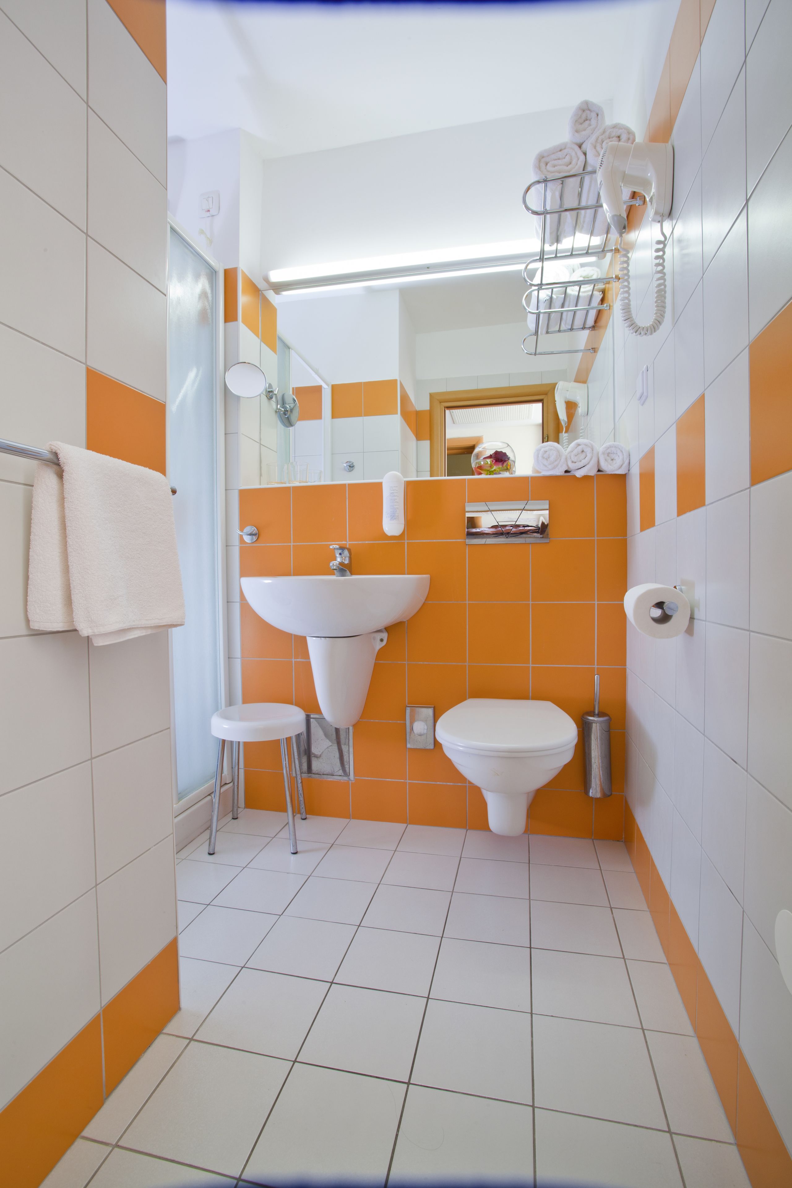 Een badkamer met een wit wastafel, toilet en een spiegel boven het wastafel. Handdoeken hangen aan een rek en een haardroger is aan de muur bevestigd.