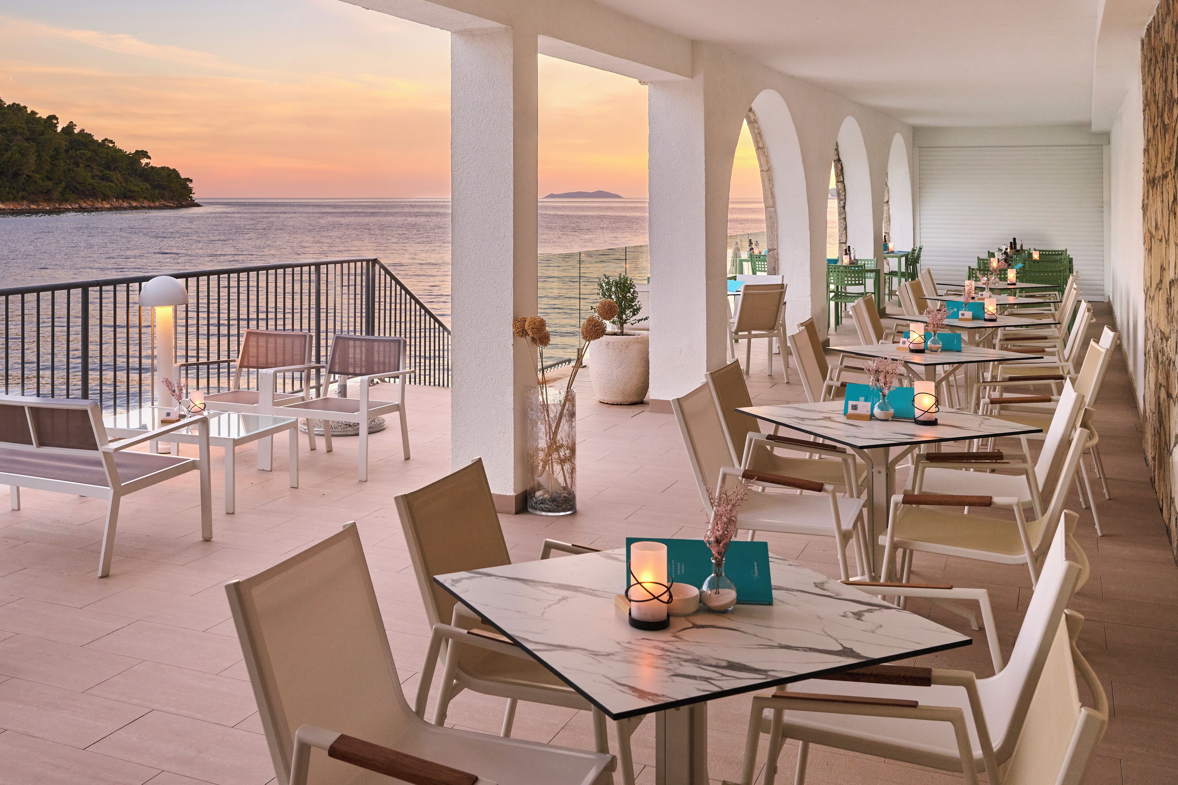 A restaurant with outdoor seating overlooking the sea, featuring white tables, chairs, and a view of the ocean.