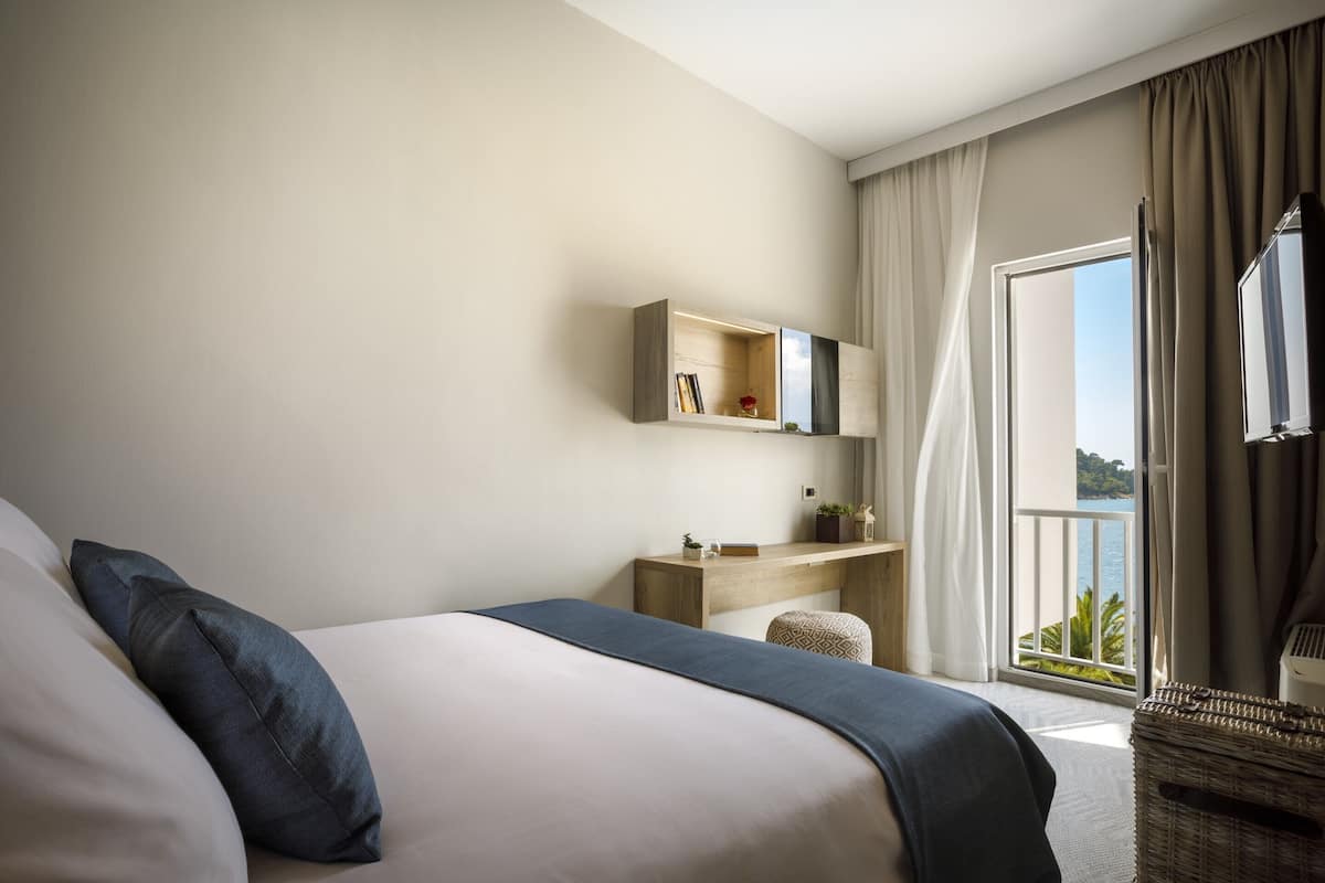 Moderni hotellihuone, jossa on suuri sänky, yöpöytä, pieni työpöytä ja parveke, jolta on näkymä.