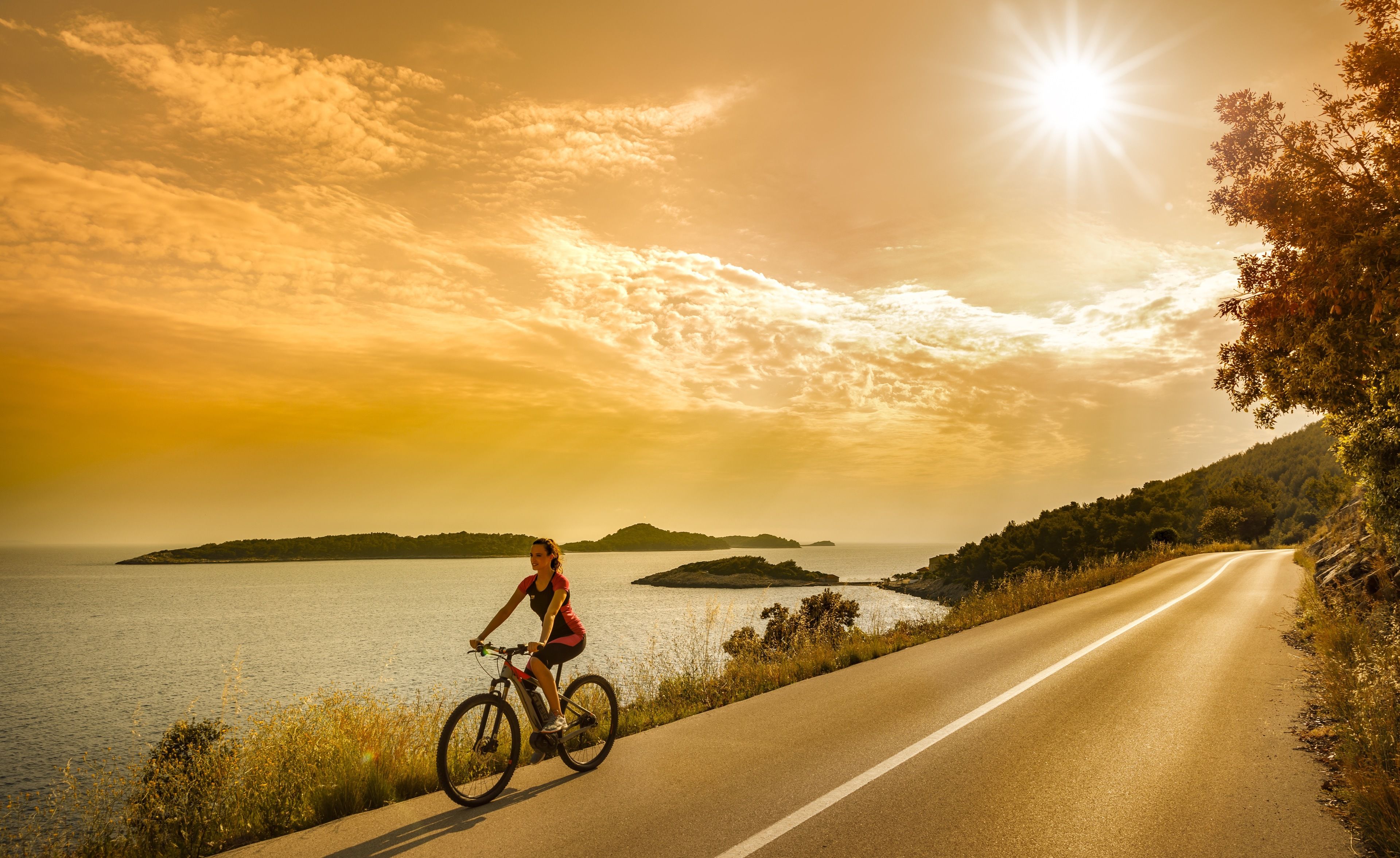 A cyclist on a paved road with a body of water and islands in the background under a sunset sky.