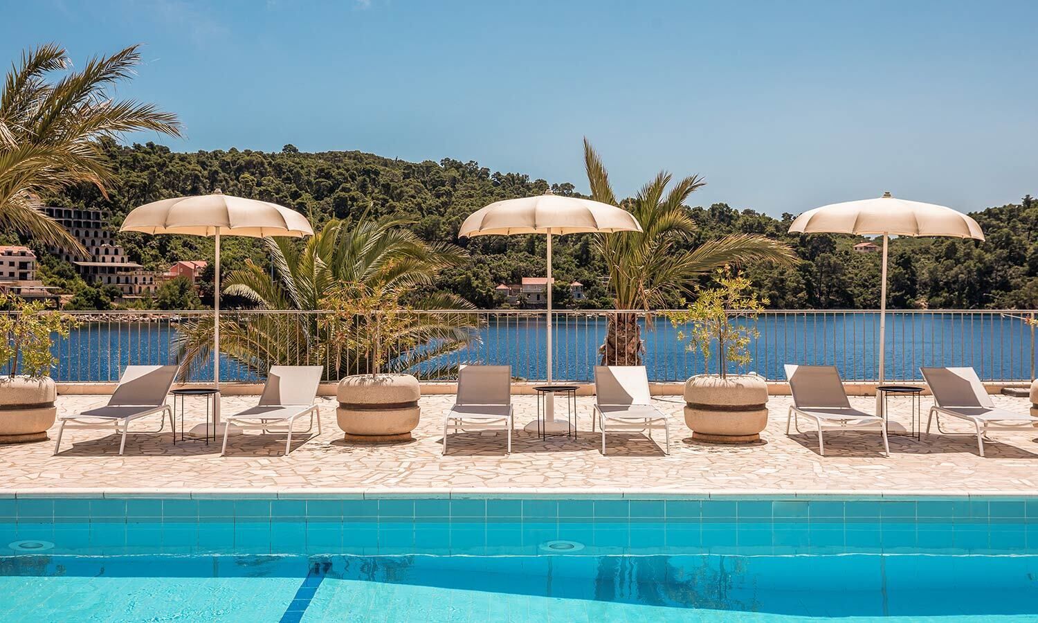 Zona junto a la piscina con sillones de descanso y sombrillas, vista despejada del agua, y arbolado con césped y valla.