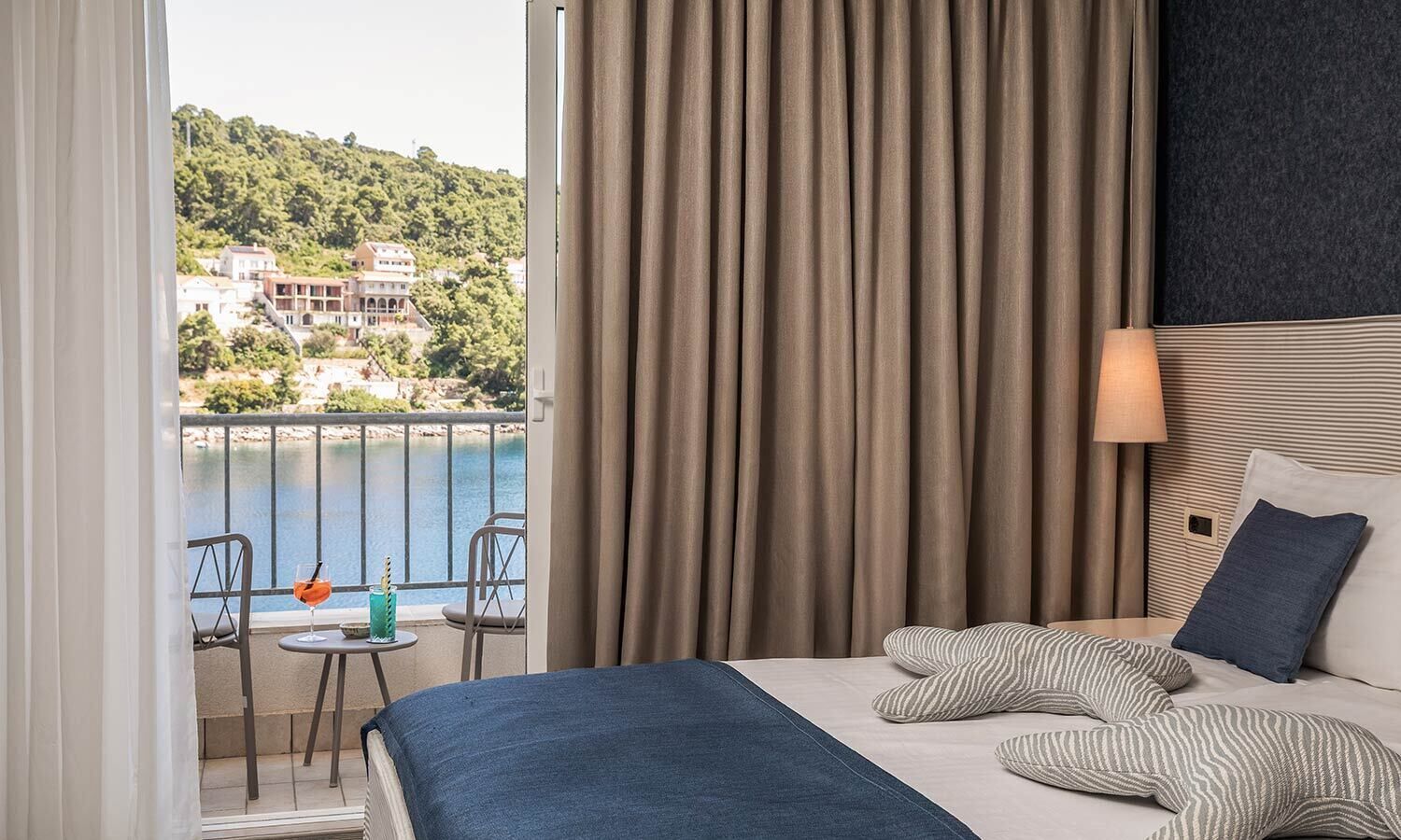 A hotel room with a balcony, a bed with striped pillows, a nightstand, and a view of a coastal area.