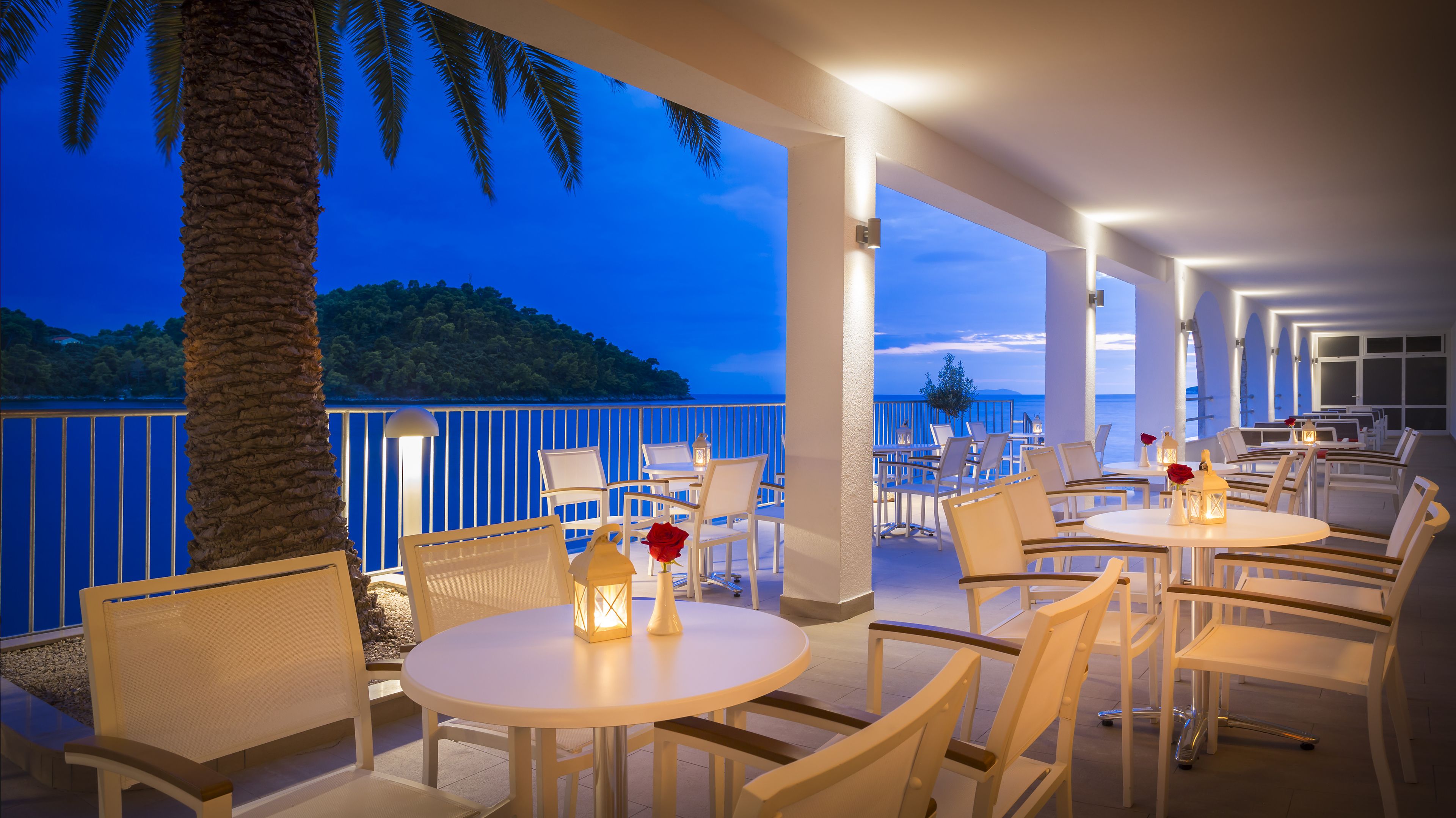A seaside restaurant with round tables and white chairs, overlooking a calm sea and a distant island.